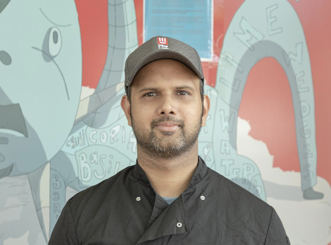 A man wearing a gray cap and black chef's coat stands in front of a colorful illustrated wall with abstract drawings and words like "water" and "basil." He is looking directly at the camera with a neutral expression.