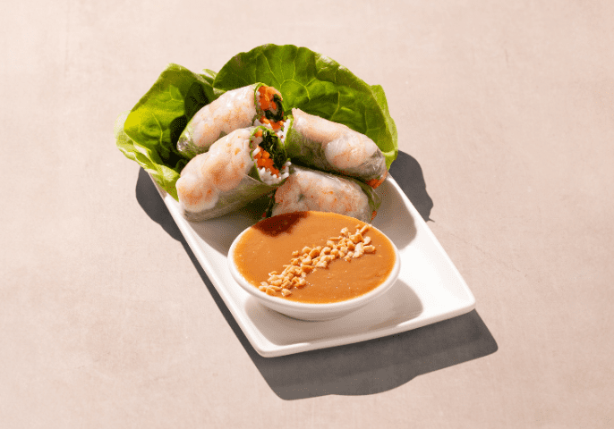 Fresh spring rolls filled with shrimp, vegetables, and herbs are neatly arranged on a white plate with lettuce. A small bowl of peanut sauce is placed beside them. The background is a smooth, light surface.