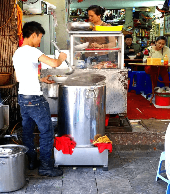 A street vendor stands by a large steaming pot, ladling soup into a bowl. Behind, a woman prepares ingredients at a small stall, while customers sit at blue tables enjoying their meals. The scene is lively, capturing the essence of street food culture.