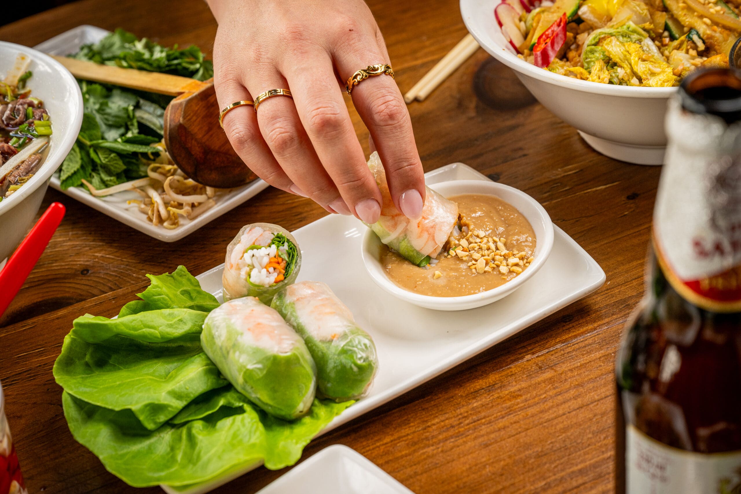 A hand dips a Vietnamese spring roll into peanut sauce on a rectangular plate, with leafy greens and more rolls. Bowls of food and a beer bottle are on a wooden table, suggesting a dining scene.