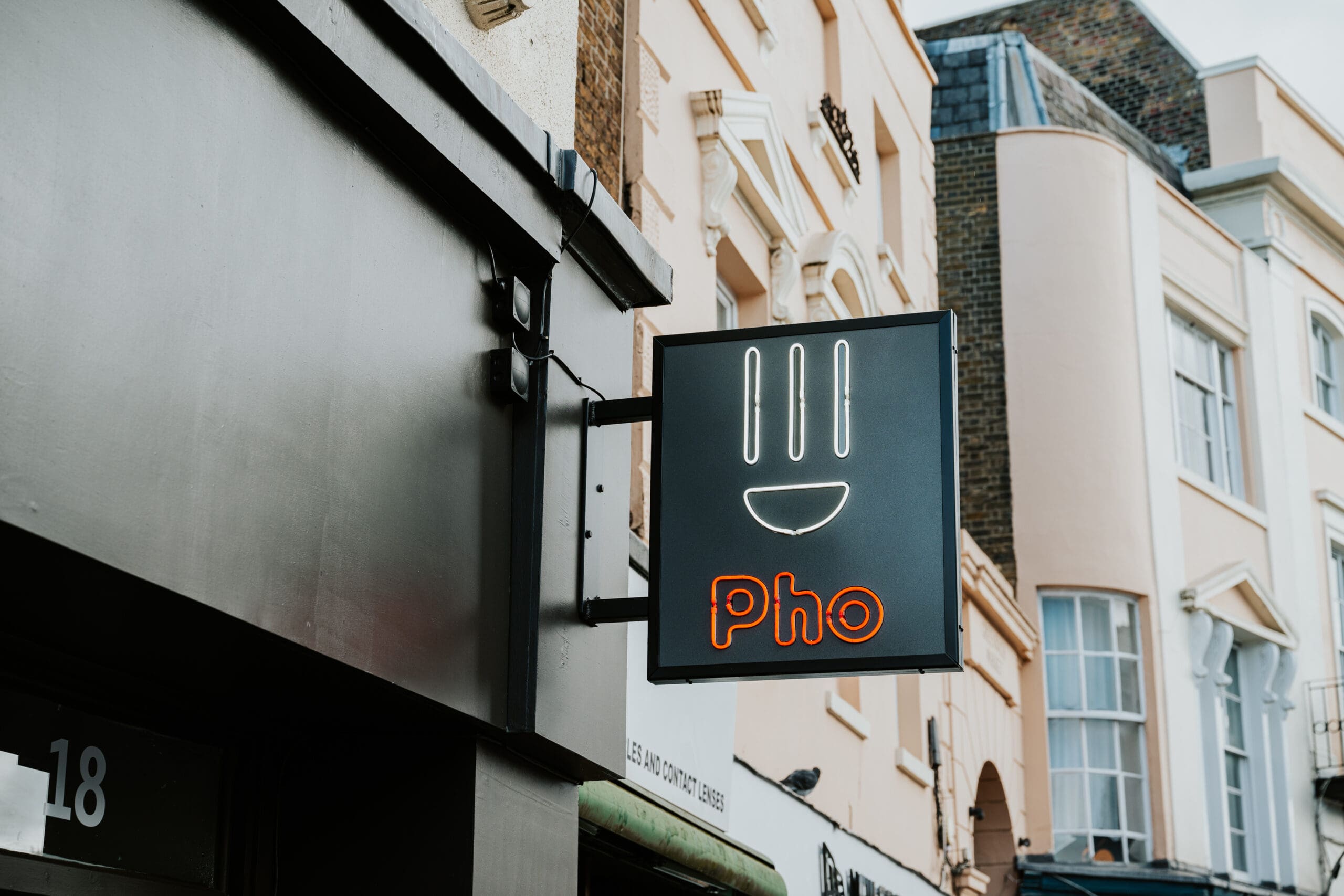 A street scene showing a square sign with a smiling face made from three vertical lines and a curved line, above the word "Pho" in red neon. The background includes several light-colored building facades.