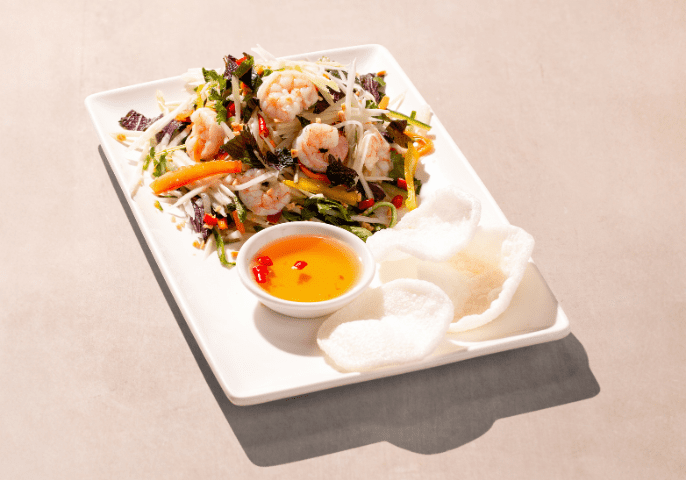A rectangular white plate with a vibrant shrimp salad, featuring mixed greens, carrots, and bean sprouts. A small dish of dipping sauce and prawn crackers are on the side. The background is a light gray surface.
