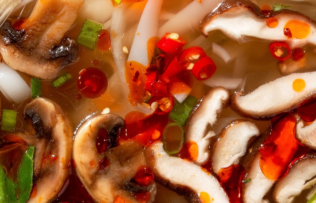 Close-up of a vibrant soup with sliced mushrooms, chopped green onions, red chili slices, and noodles in a savory broth. Red oil droplets float on the surface, adding a touch of spice.