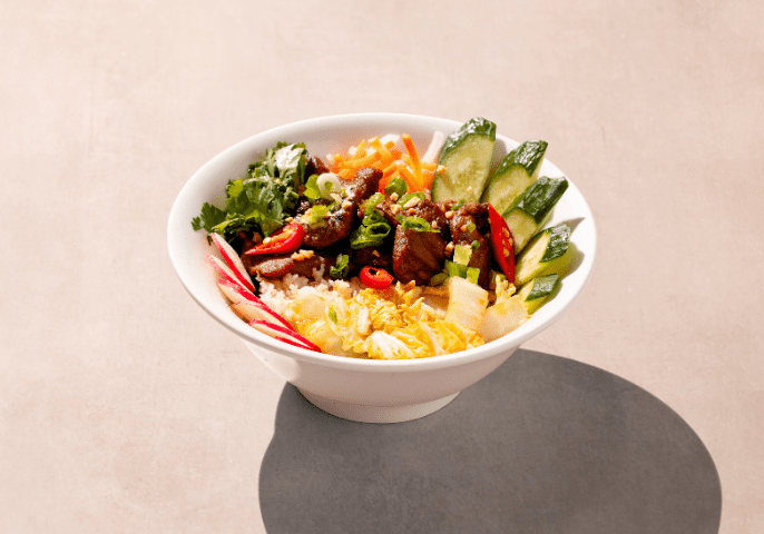 A white bowl filled with rice, topped with sliced cucumbers, cilantro, thin radish slices, red peppers, shredded carrots, green onions, and a mix of cooked beef pieces. The background is a neutral, light surface.