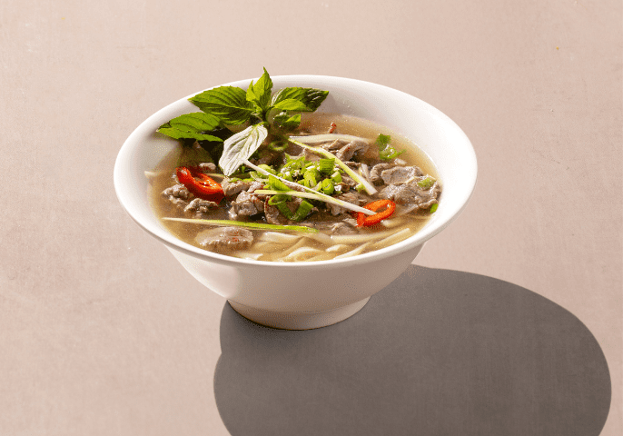 A white bowl of pho featuring broth, noodles, beef slices, fresh herbs, red chili slices, and bean sprouts, set on a neutral background.