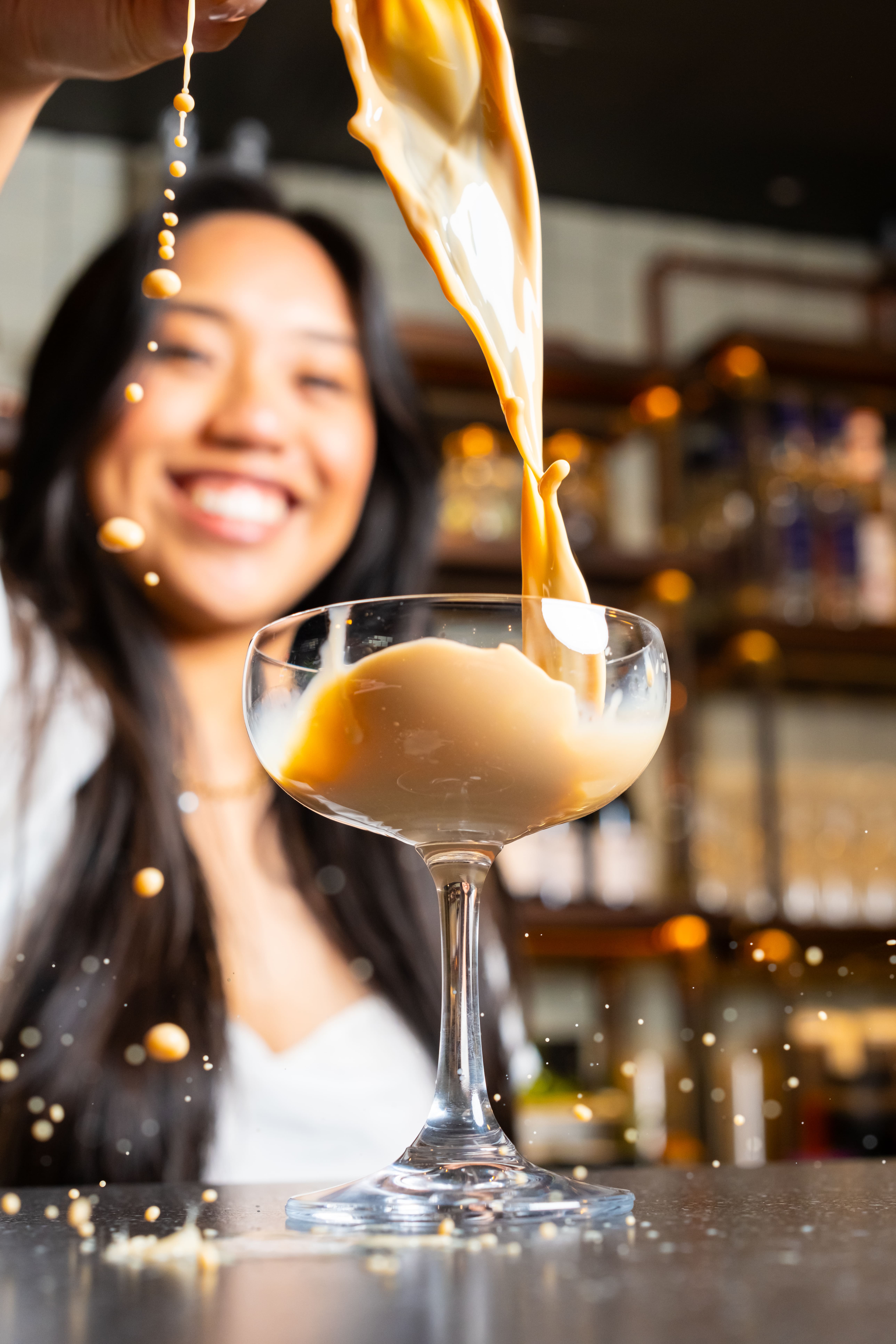 A woman joyfully pours a creamy, coffee-colored liquid into a cocktail glass, with splashes captured mid-air. She stands in a warmly lit setting, possibly a bar or cafe, with shelves in the background.