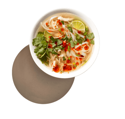 A bowl of Vietnamese pho with noodles, chicken slices, cilantro, lime, green onions, and red chili peppers in broth, viewed from above. The bowl is placed on a wooden surface.