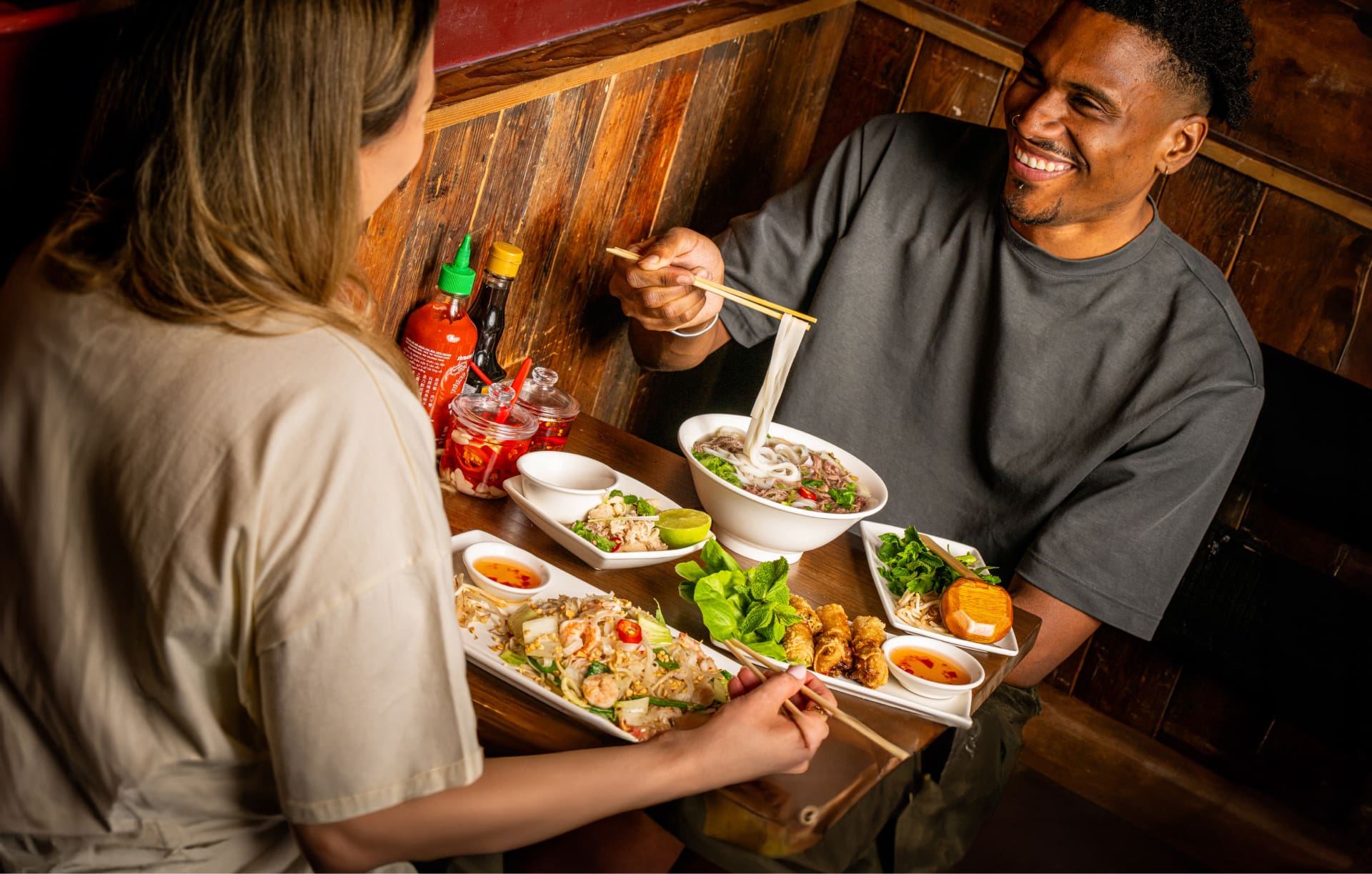 A man and woman are sitting at a wooden table, smiling and enjoying a meal. They have dishes of noodles and vegetables in front of them, along with sauces. Various condiments are on the table, and the setting is warmly lit.