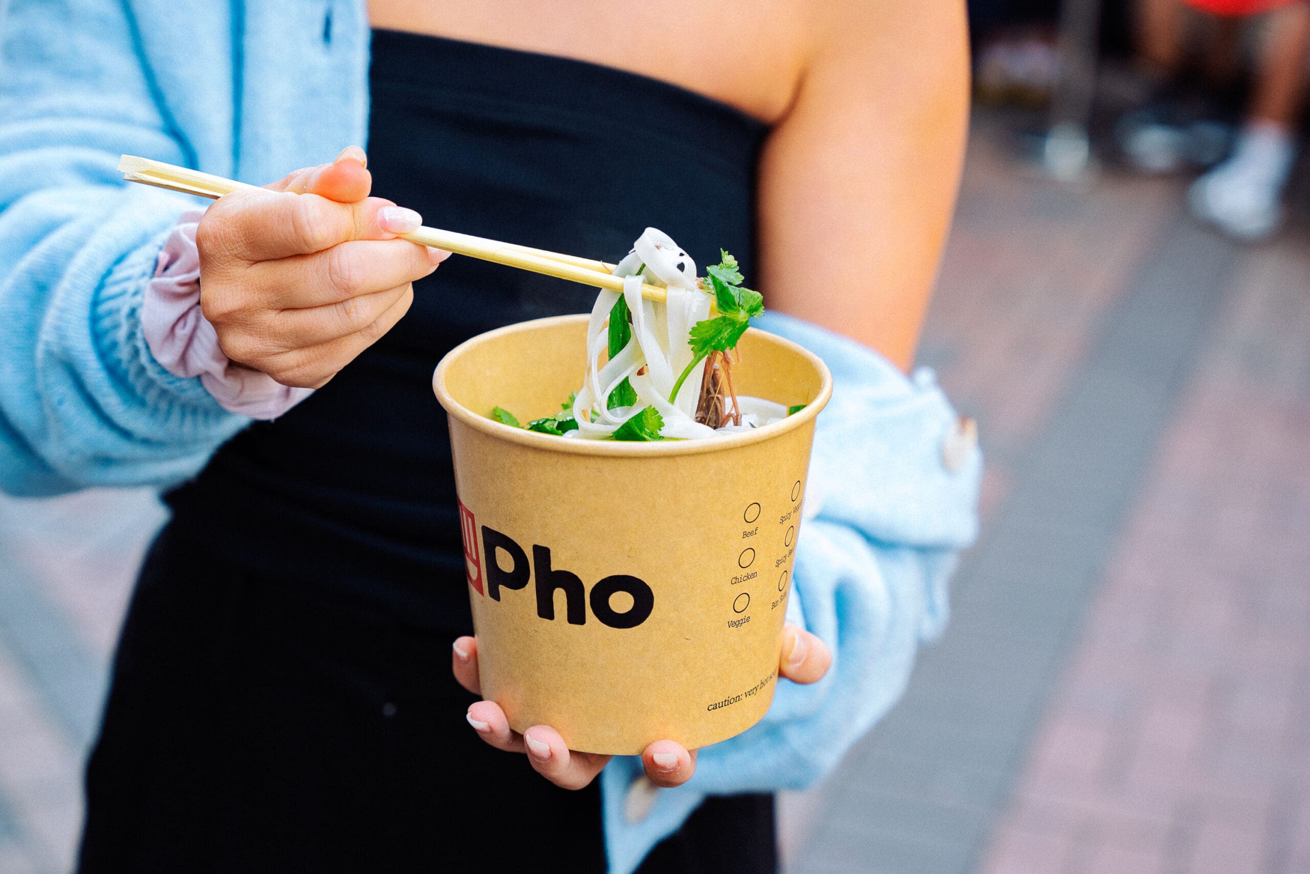 A person in a black top and blue sweater holds a cardboard cup filled with pho noodles, using chopsticks to lift the noodles. The cup is branded with the word "pho." The background is a blurred outdoor setting.