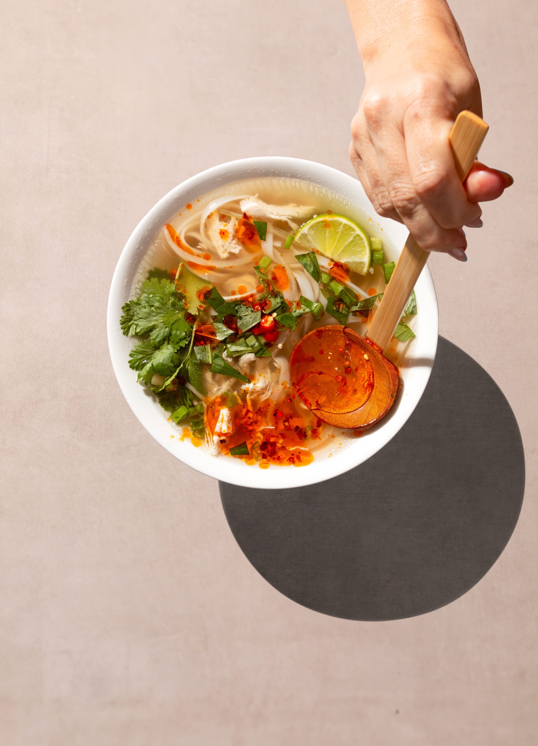 A hand holding a wooden ladle above a bowl of soup filled with noodles, herbs, lime slices, and topped with red pepper flakes. The bowl casts a shadow on a light beige surface.