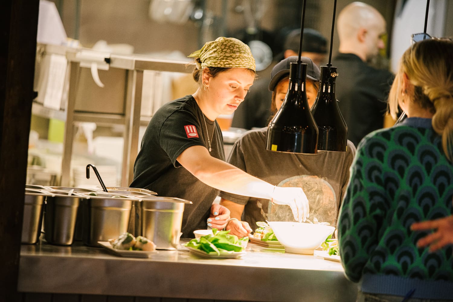 A chef wearing a headscarf and black shirt is plating food in a restaurant kitchen, illuminated by hanging lights. Another person assists beside her. Tubs and prepared ingredients are on the counter.