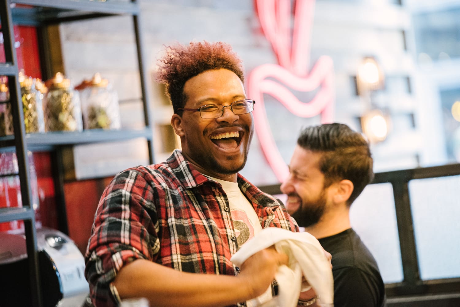 Two men laughing in a brightly lit cafe. One wears glasses and a red flannel shirt, holding a cloth. The other has a beard and wears a black shirt. Shelves with jars and a neon sign are in the blurred background.