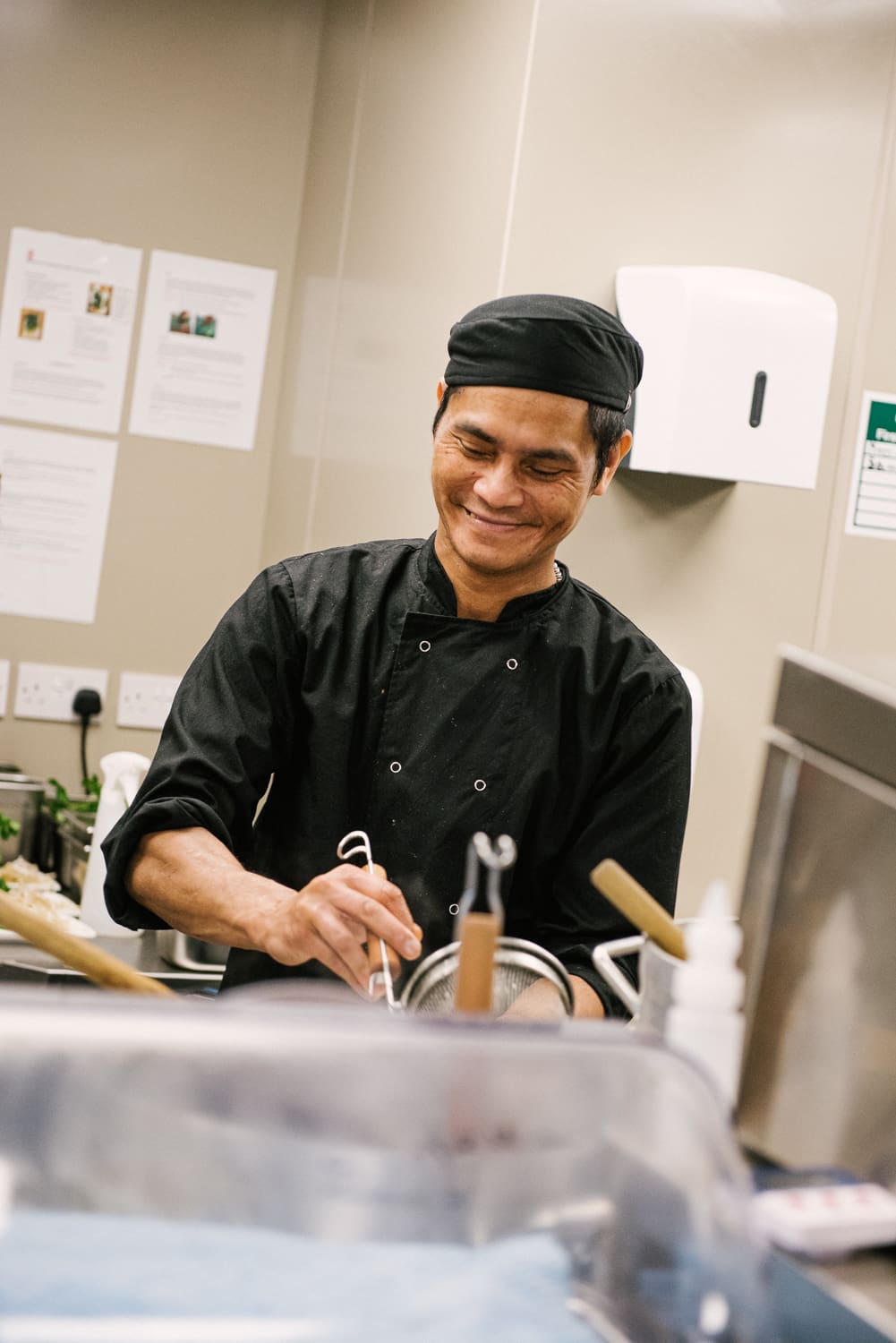 A chef in a black uniform and hat smiles while preparing food in a kitchen. He stands near a countertop with various kitchen utensils visible. The background shows a beige wall with papers and a dispenser.