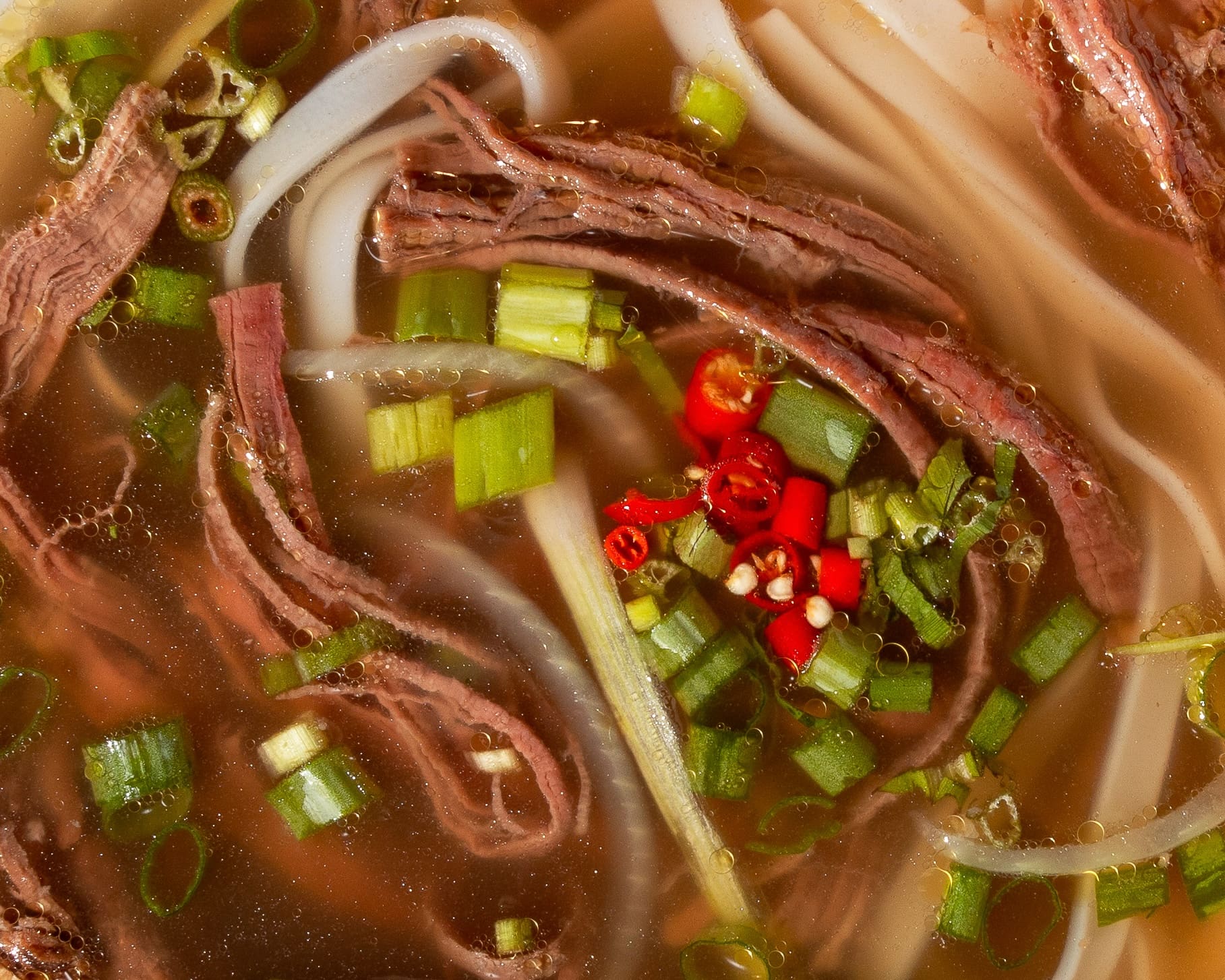 Close-up of a bowl of noodle soup with sliced beef, red chili peppers, green onions, and noodles in a rich broth. The ingredients are arranged in a visually appealing manner, showcasing vibrant colors and textures.
