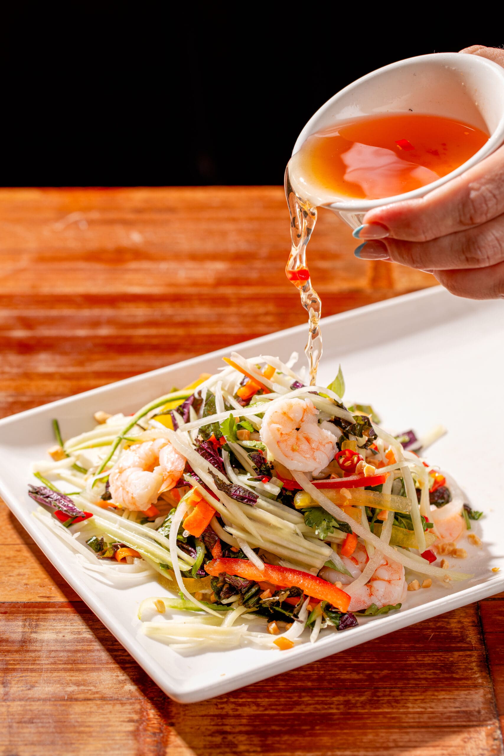 A hand pours dressing over a vibrant salad with sliced shrimp, green papaya, carrots, and other colorful vegetables, served on a white rectangular plate on a wooden table.