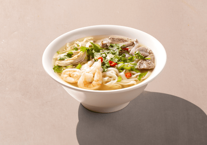 A bowl of noodle soup with shrimp, sliced fish, green herbs, and red chili slices, served in a white bowl on a light surface.