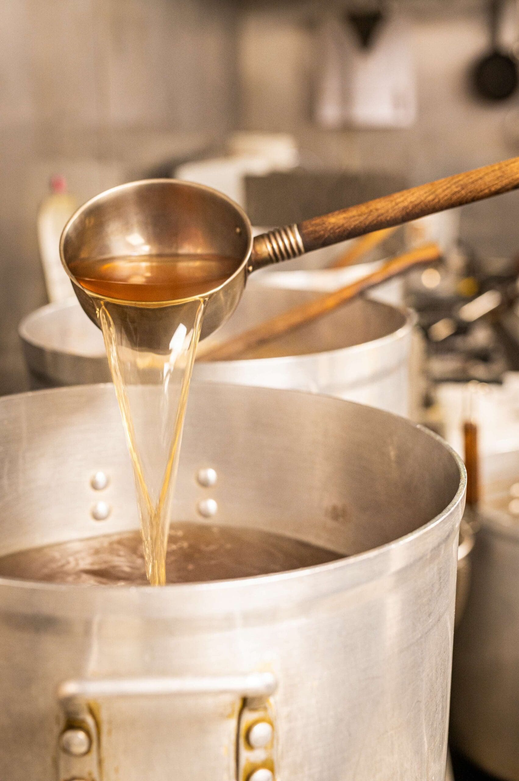 A ladle pours broth into a large metal pot in a kitchen setting. The broth has a warm, light brown color, and steam rises from the pot, suggesting warmth and a cooking process underway. The background is slightly blurred.