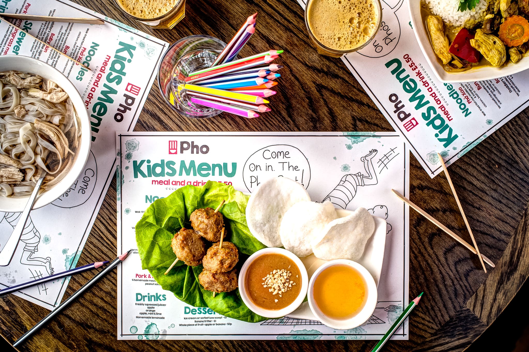 Kids menu placemat with coloring pencils, noodle soup, lettuce wraps with dipping sauces, and drinks on a wooden table. The placemat has playful drawings and text inviting coloring activities.