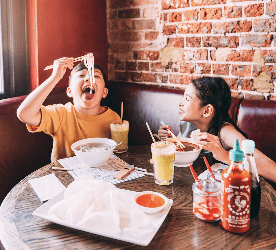 Two children enjoying noodles at a restaurant. The boy is playfully lifting noodles with chopsticks, while the girl smiles with a bowl in front of her. The table is filled with food, drinks, and condiments, set against a brick wall.