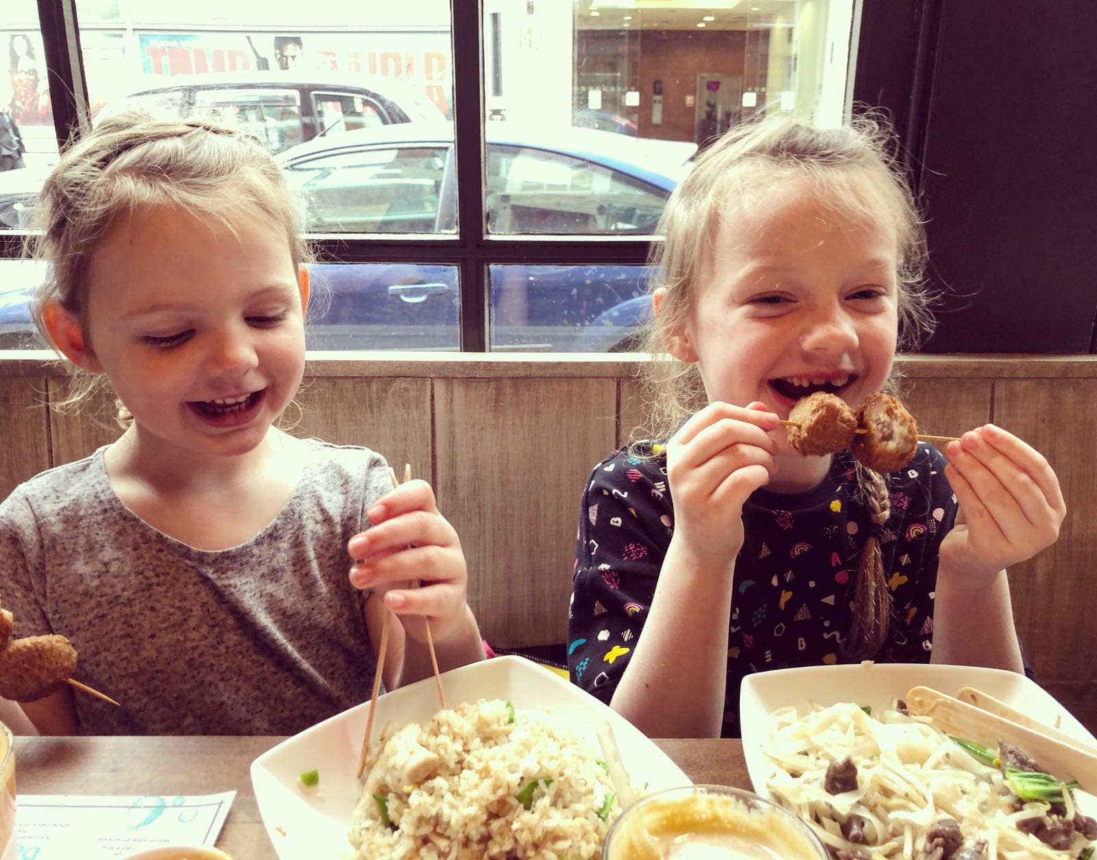 Two young girls sit at a table in a restaurant, smiling and laughing while eating skewered food over bowls of noodles and rice. A window behind them shows cars parked outside on the street.