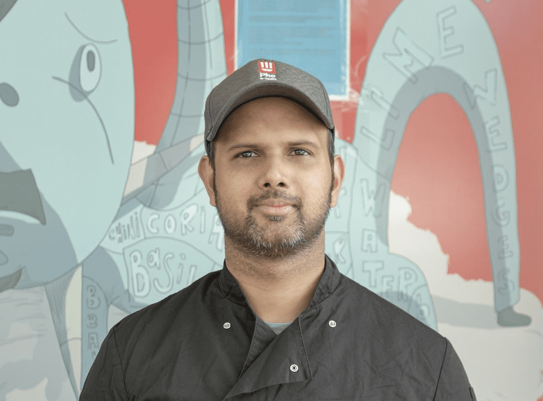 A man wearing a gray cap and black chef's coat stands in front of a colorful illustrated wall with abstract drawings and words like "water" and "basil." He is looking directly at the camera with a neutral expression.