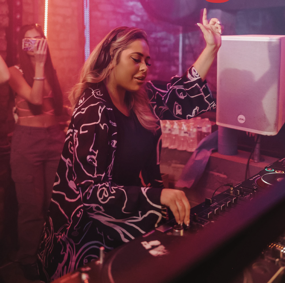 A DJ wearing headphones and a patterned jacket is mixing music at a DJ booth in a dimly lit club, raising one hand. A person in the background records the scene with a phone. Neon pink lights create a vibrant atmosphere.