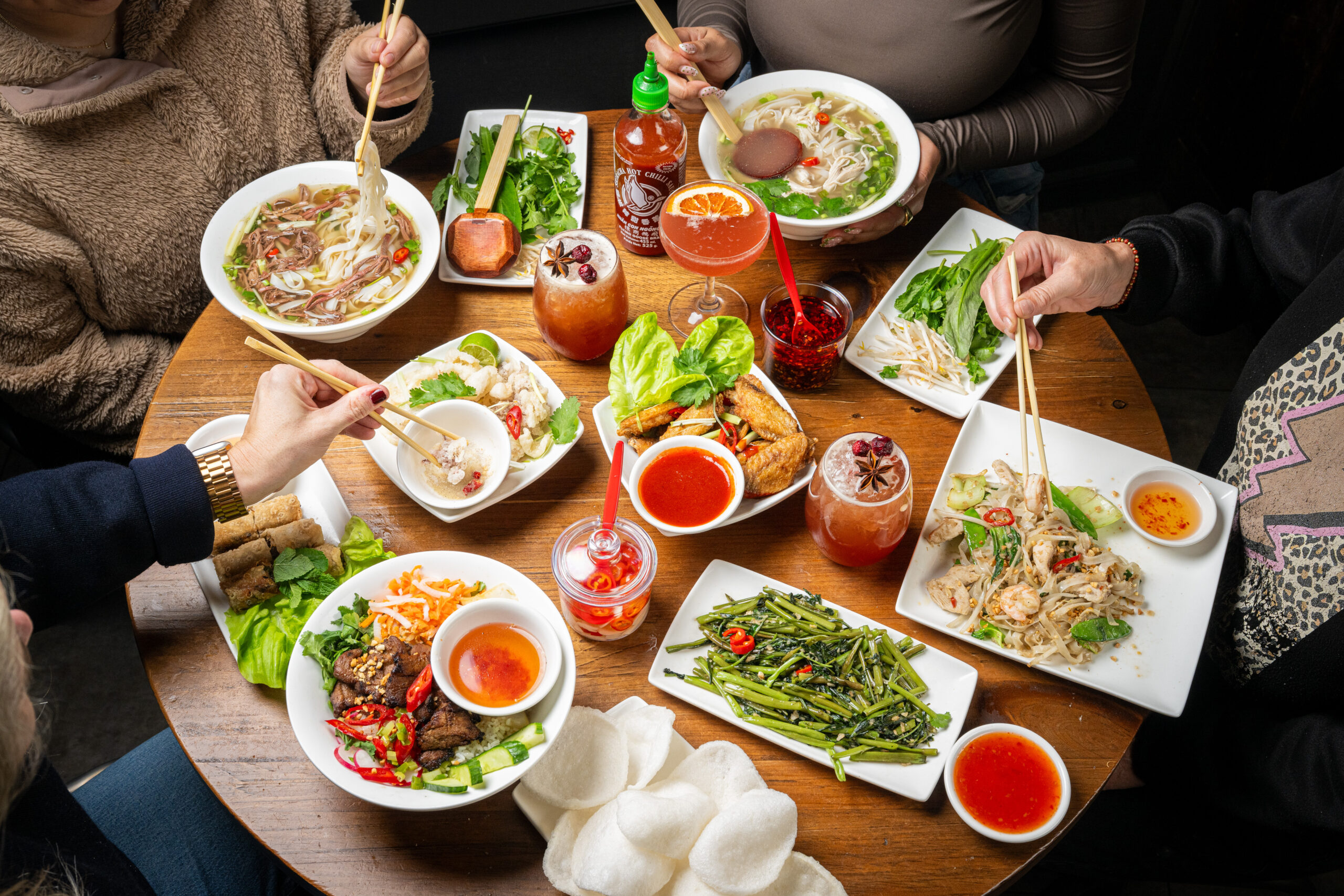 Four people sit around a wooden table filled with Vietnamese dishes, including pho, spring rolls, dumplings, grilled meat, sautéed greens, dipping sauces, and colorful drinks. Hands use chopsticks to serve and eat.
