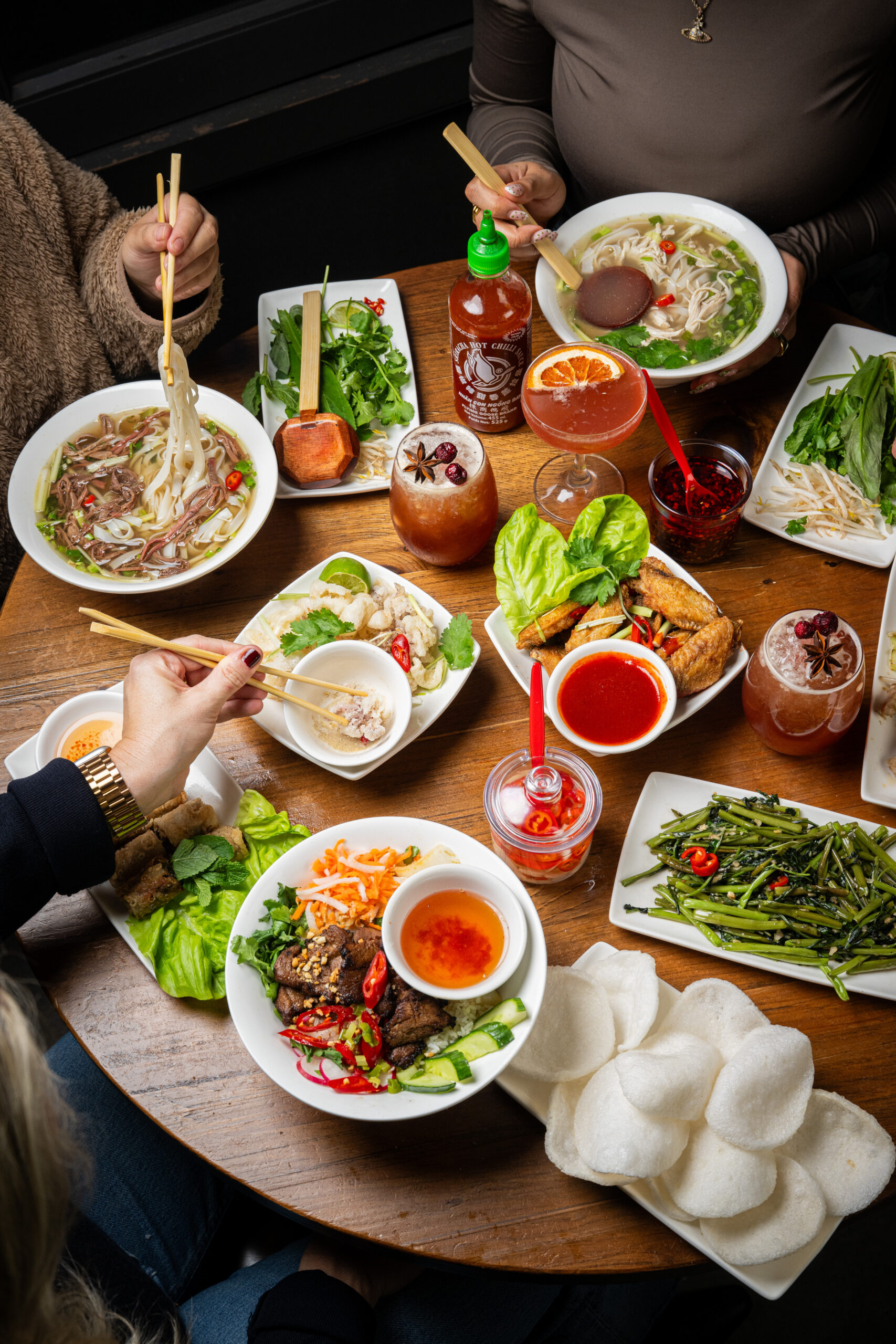 A table filled with Vietnamese dishes, including pho, rice bowls, spring rolls, fresh herbs, sauces, green beans, rice crackers, and drinks. Three people are reaching for food with chopsticks and forks.