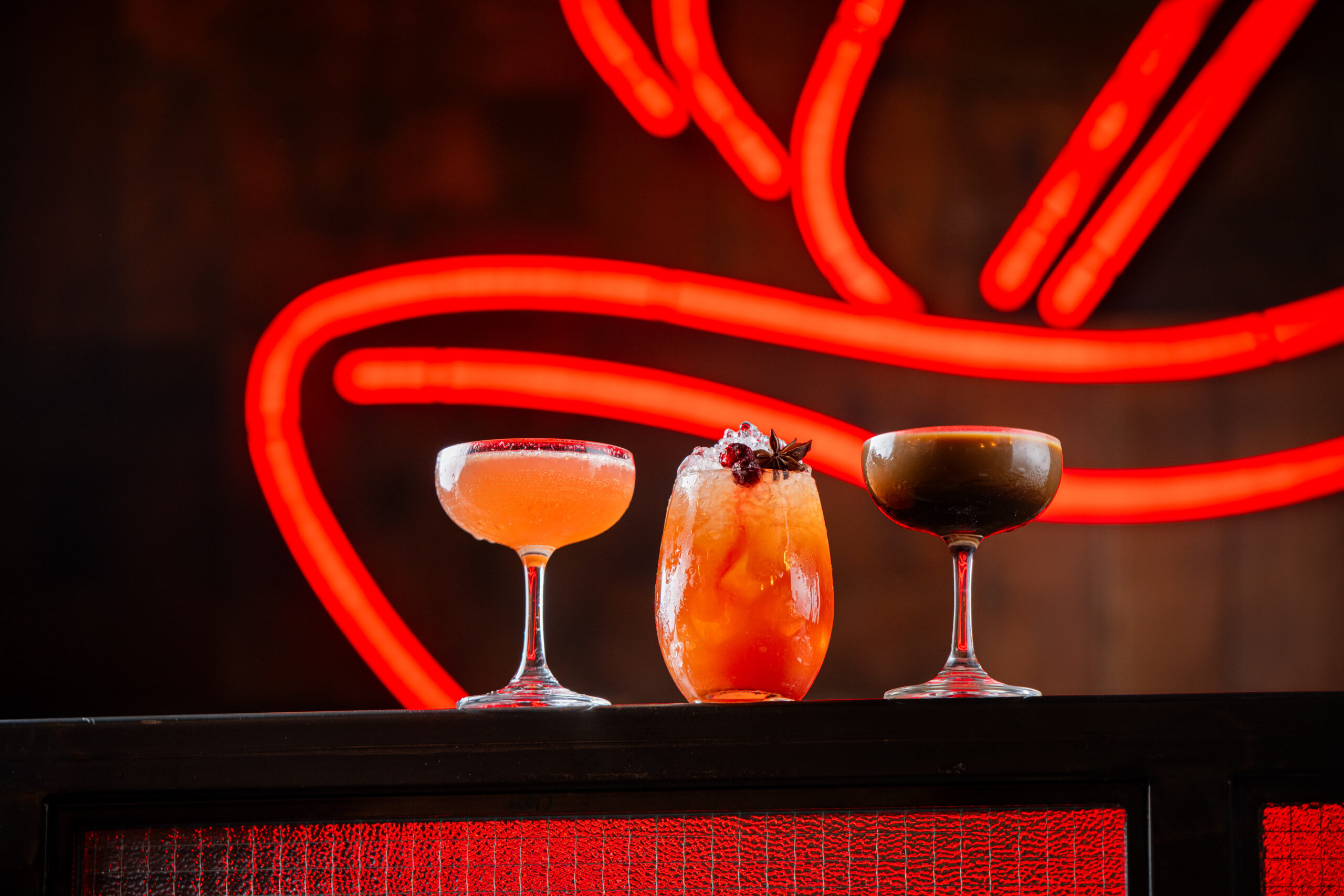 Three colorful cocktails in elegant glasses are arranged on a black surface, with a bright red neon sign glowing in the blurred background.