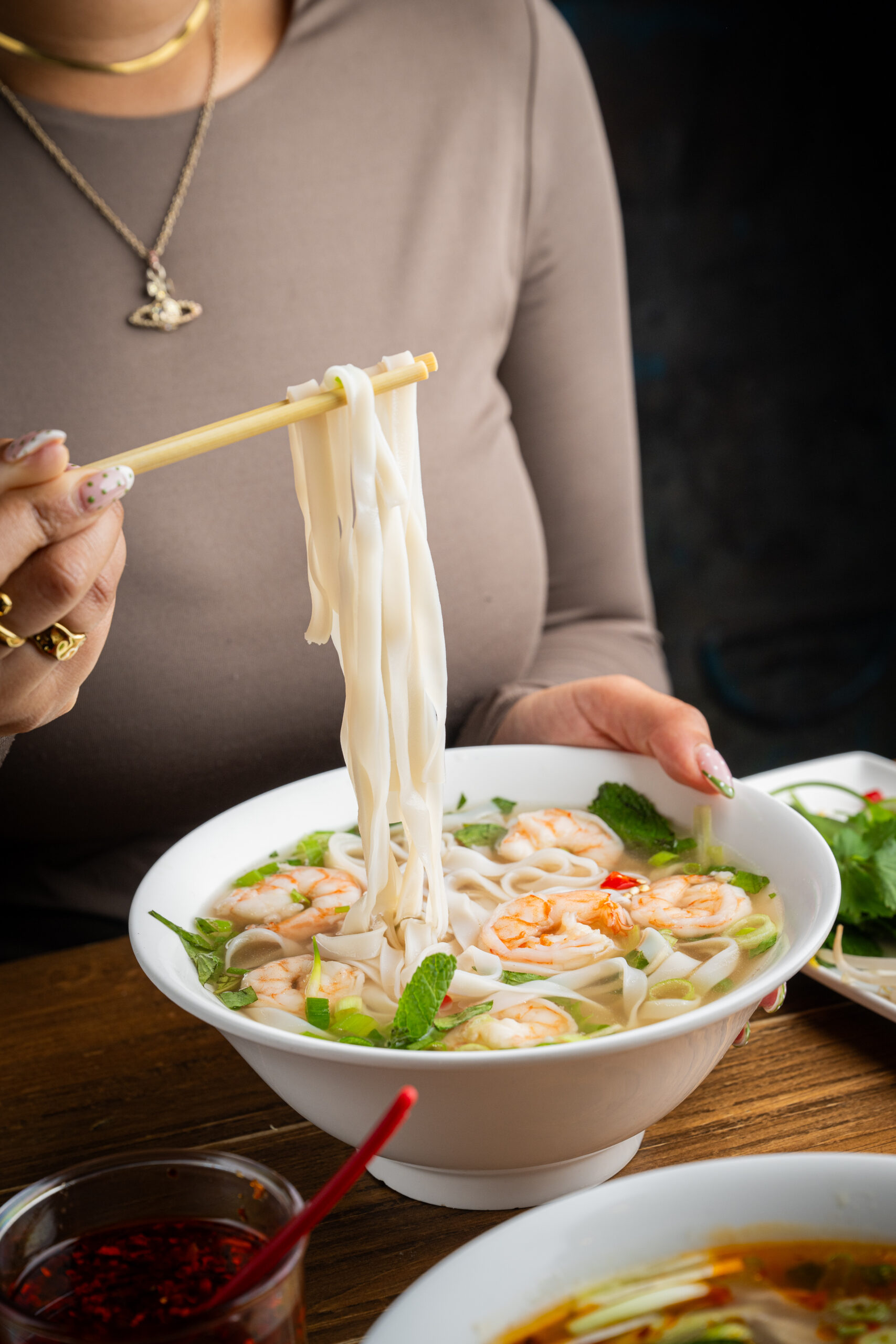 A person holding chopsticks lifts rice noodles from a bowl of Vietnamese pho, garnished with shrimp, herbs, and vegetables. Another bowl of soup and a condiment dish are on the table.
