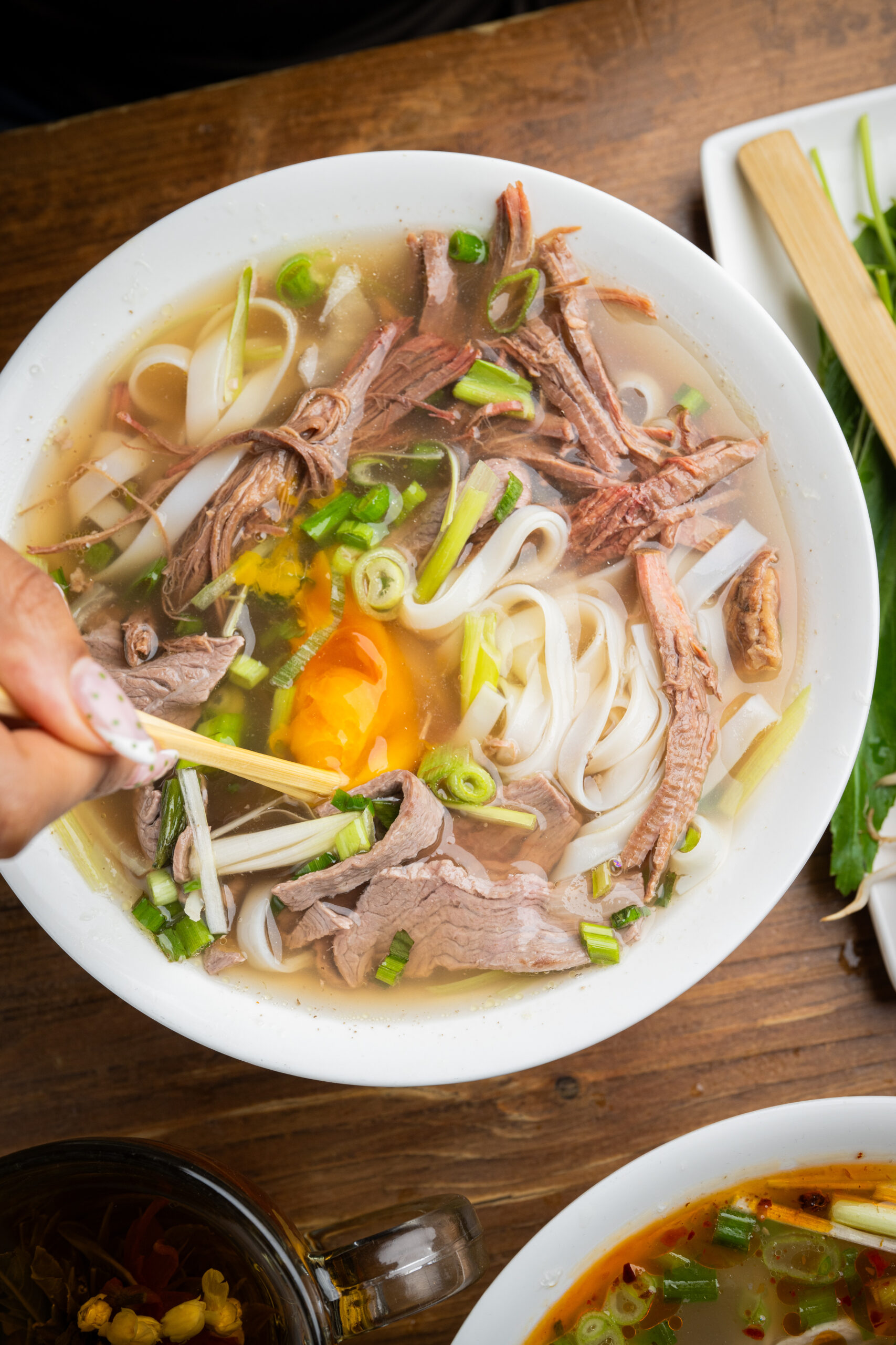 A hand uses chopsticks to mix a bowl of Vietnamese pho with rice noodles, slices of beef, green onions, and a raw egg yolk in clear broth, on a wooden table with herbs and soup nearby.