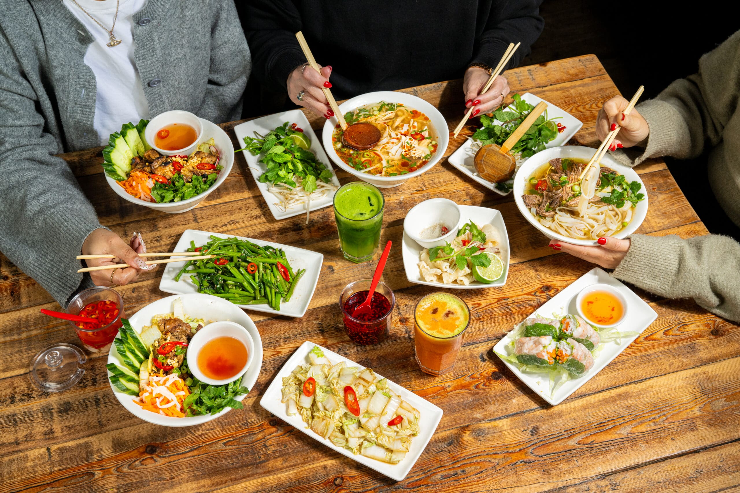 Three people sit at a wooden table sharing a colorful Vietnamese meal with noodle bowls, fresh herbs, spring rolls, grilled vegetables, dipping sauces, and drinks, using chopsticks to eat.