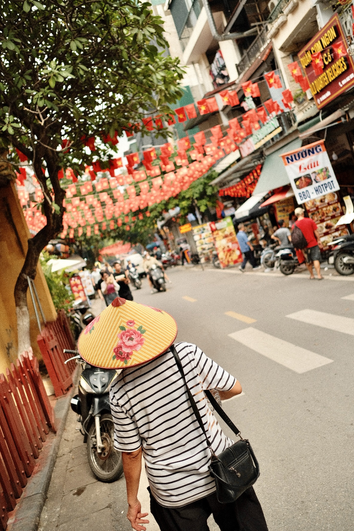 A person wearing a conical hat with a floral design and striped shirt walks down a lively street decorated with red flags and lanterns, surrounded by shops, signs, motorbikes, and trees.