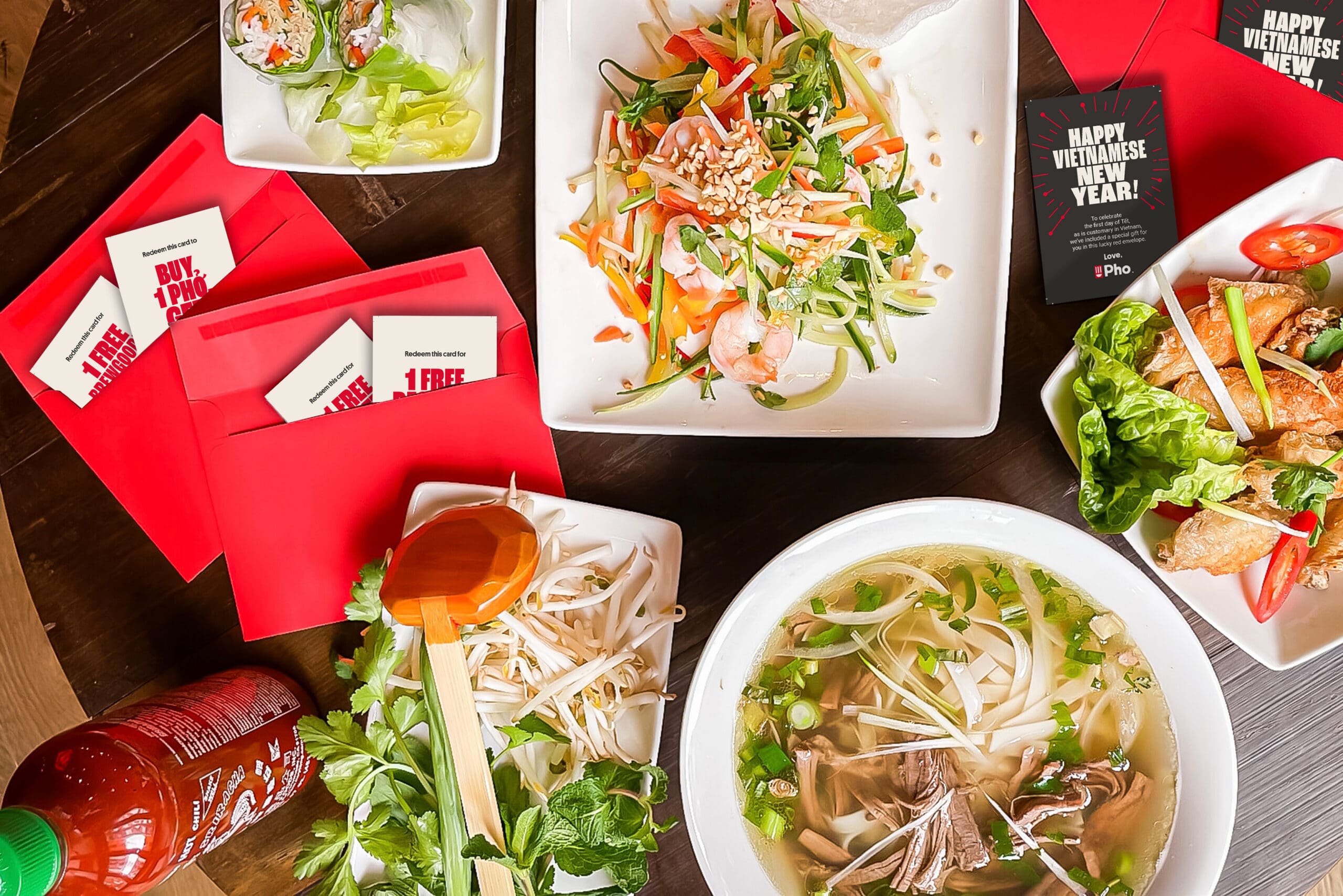 A Vietnamese meal with pho, spring rolls, salad, fresh herbs, Sriracha, and red envelopes containing coupons, alongside cards wishing “Happy Vietnamese New Year!” displayed on a wooden table.