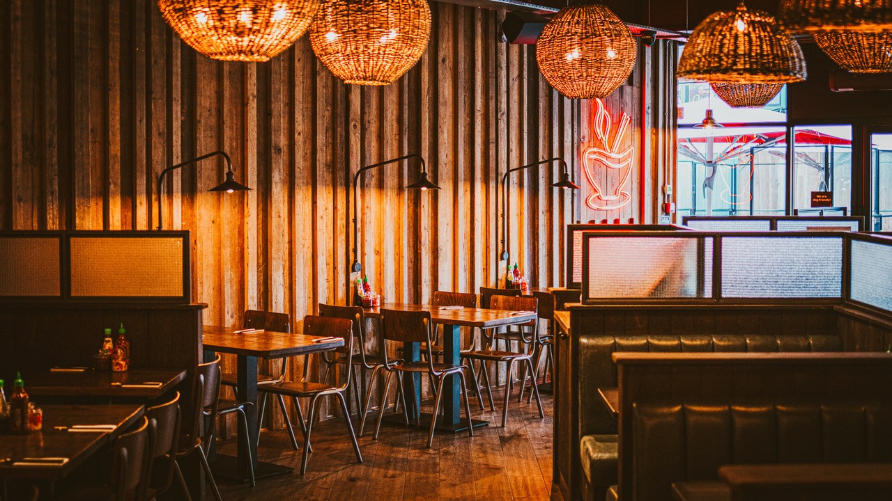 Cozy restaurant interior with wooden walls, hanging woven light fixtures, empty tables and chairs, and booths. Condiment bottles are on the tables, and a red neon sign shaped like a bowl is on the wall.