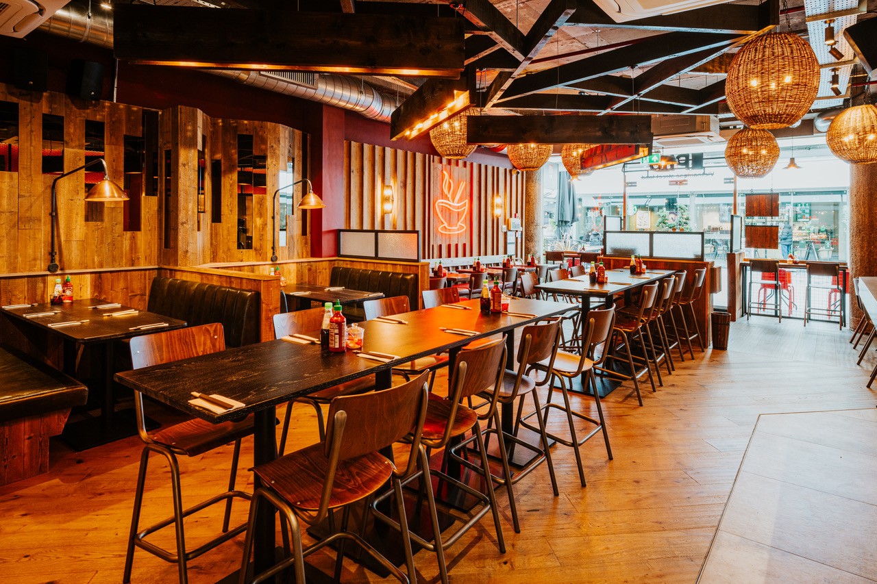 A modern restaurant interior with wooden tables and chairs, warm lighting, hanging wicker lamps, and a neon coffee cup sign on a wooden-paneled wall, creating a cozy and inviting atmosphere.