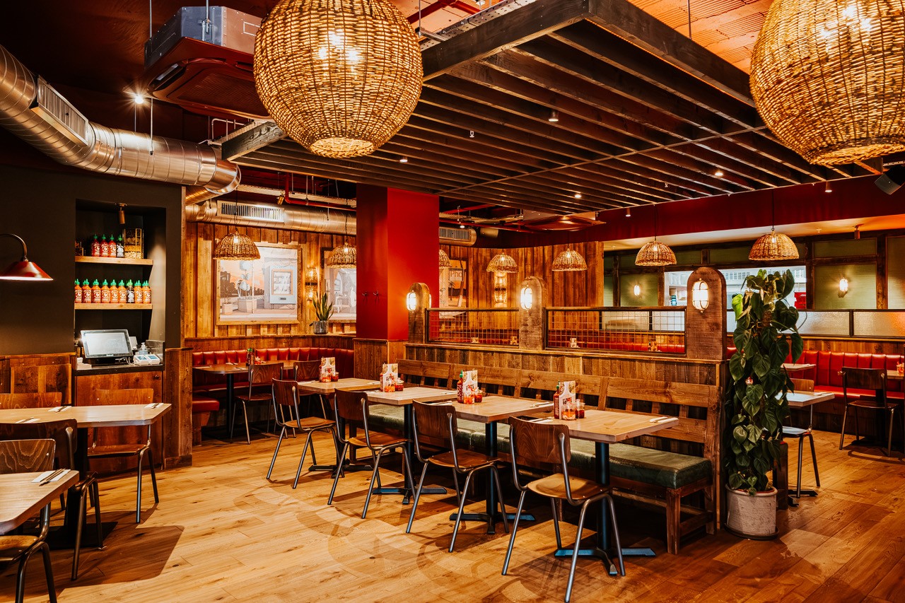 A cozy restaurant interior with wooden furniture, warm lighting from large woven pendant lamps, red accents, and plants. Booth and table seating are visible, and shelves with condiments line one wall. The atmosphere is inviting and rustic.