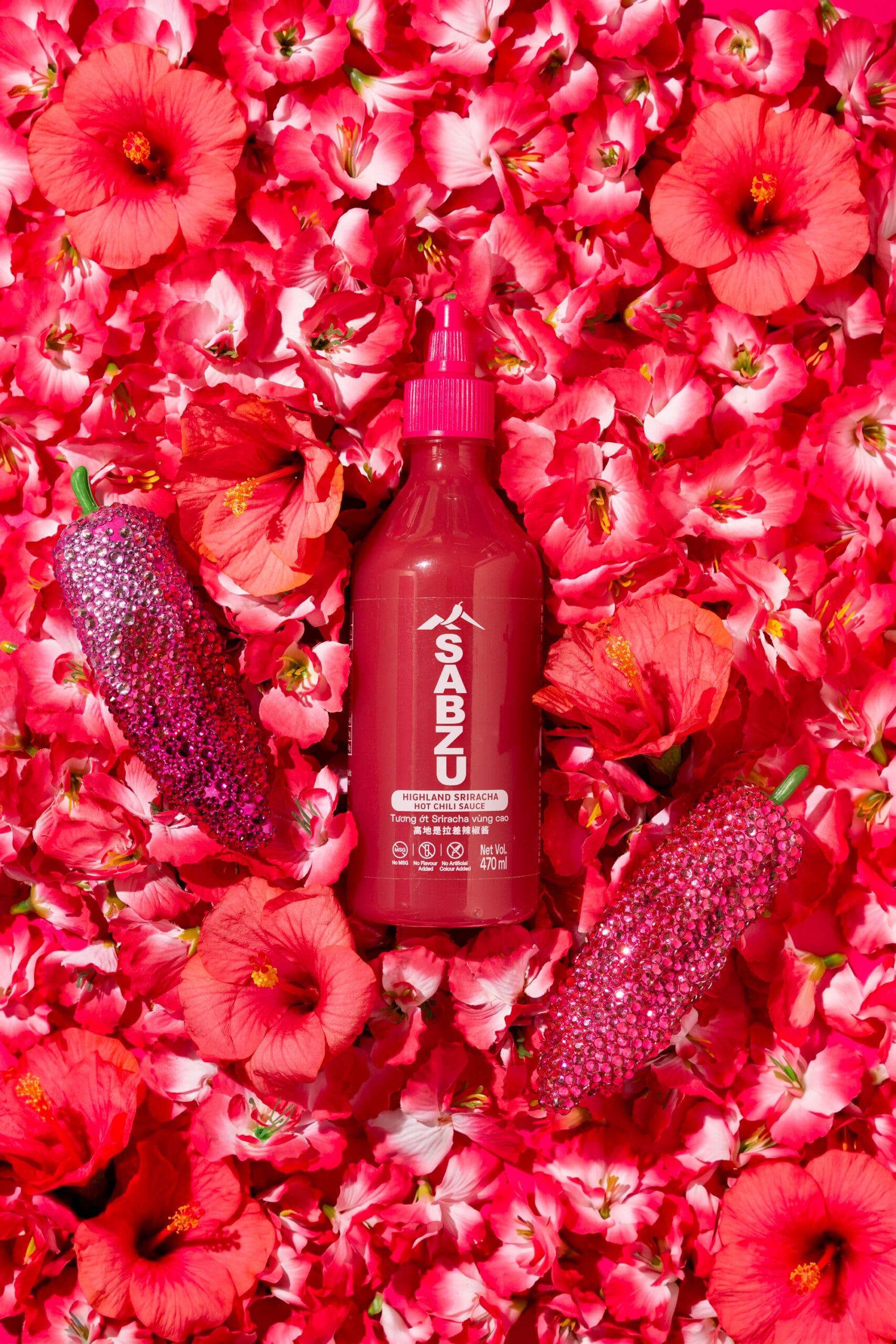 A red bottle labeled "SABZU" stands upright on a bed of bright pink hibiscus flowers, flanked by two glittery red chili peppers.
