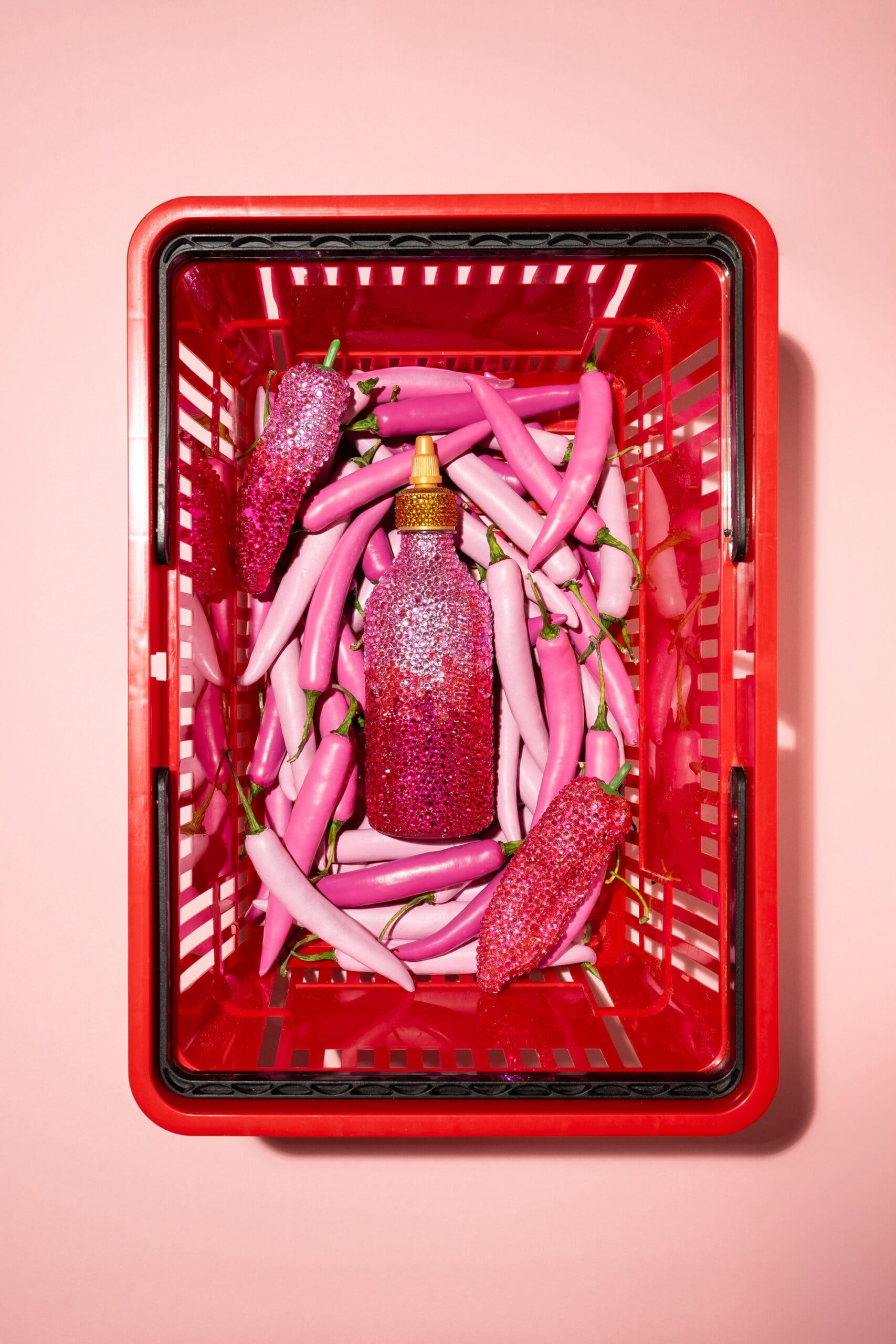 A red shopping basket filled with pink chili peppers and a pink glitter-covered hot sauce bottle, all set against a pink background.