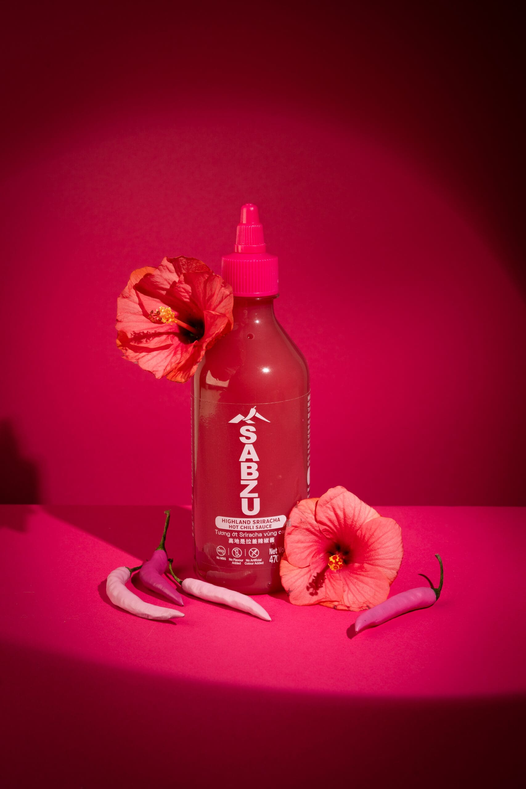 A bottle of Sabzu sauce with a pink cap is surrounded by pink chili peppers and hibiscus flowers, all set against a vibrant pink background.
