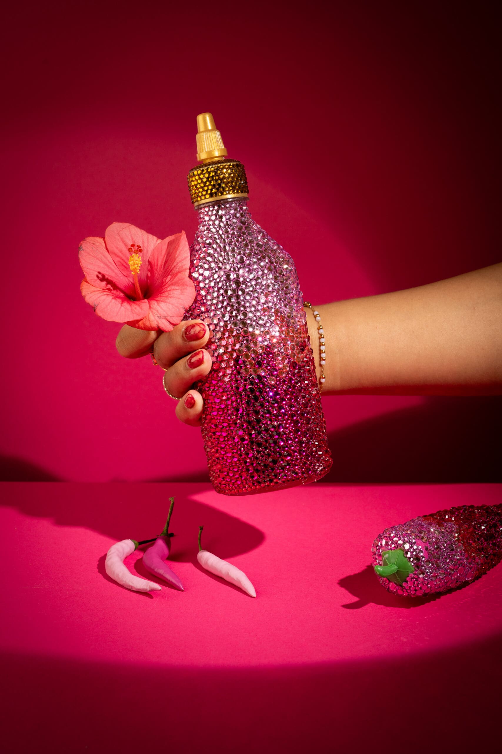 A hand holding a sparkling pink rhinestone-covered squeeze bottle with a gold cap and a pink hibiscus flower, set against a vibrant pink background. Nearby are pink chili peppers and another glittery bottle.