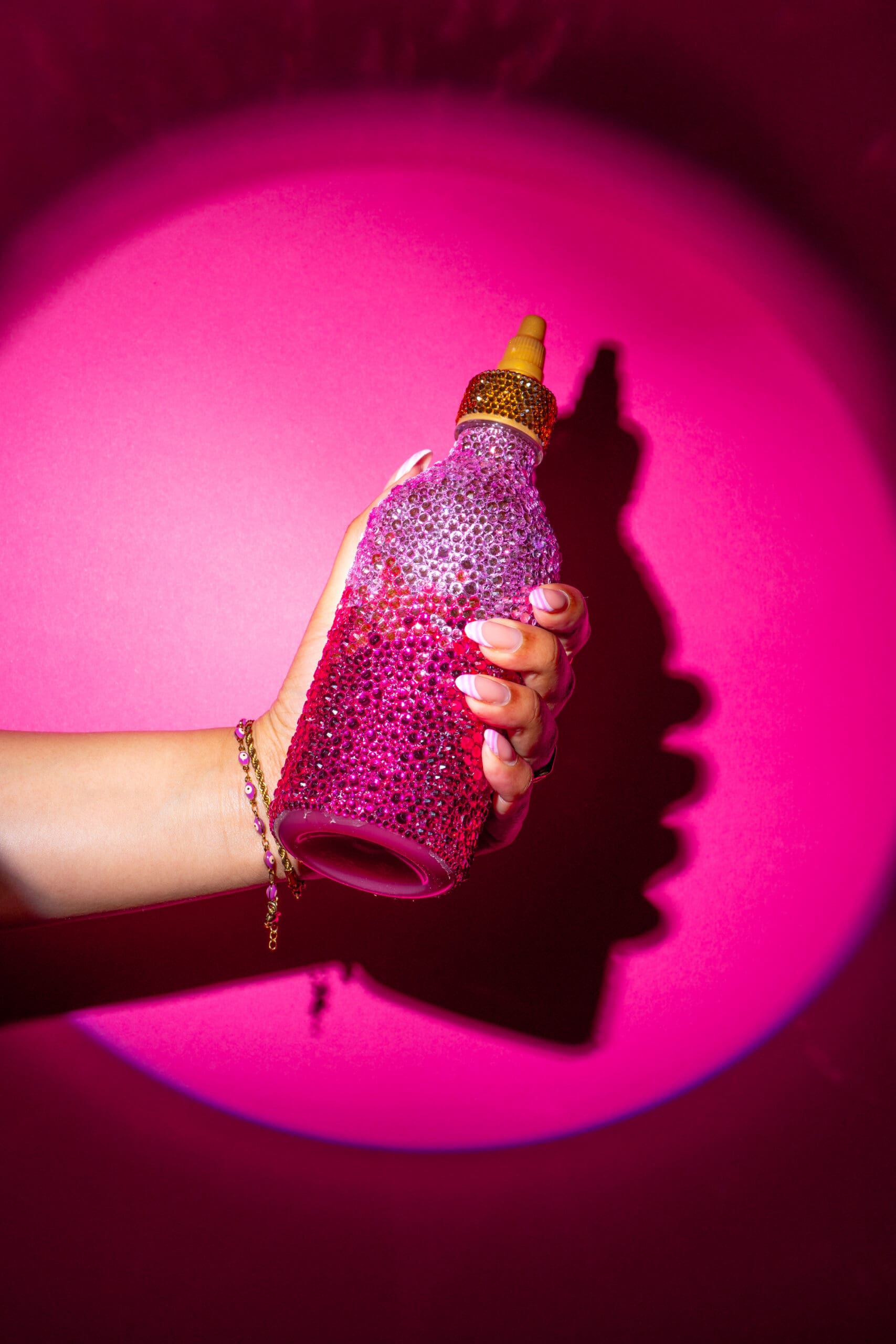 A hand with manicured nails and bracelets holds a bottle covered in pink and gold rhinestones, with a yellow nozzle, against a vibrant pink background with dramatic lighting and shadow.
