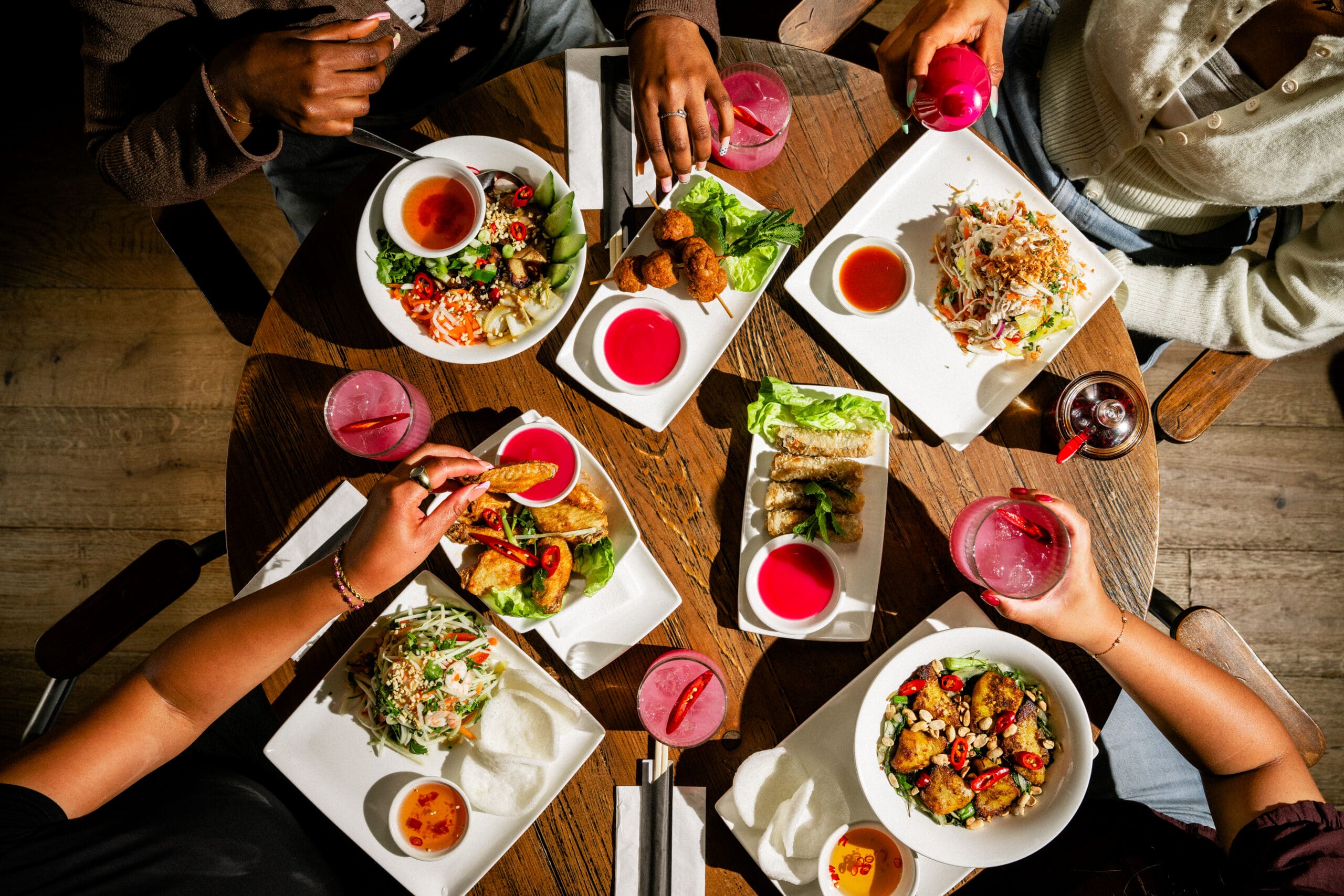 Four people sit around a wooden table enjoying colorful plates of Asian cuisine, including salads, noodles, spring rolls, and pink drinks, with chopsticks and dipping sauces visible.