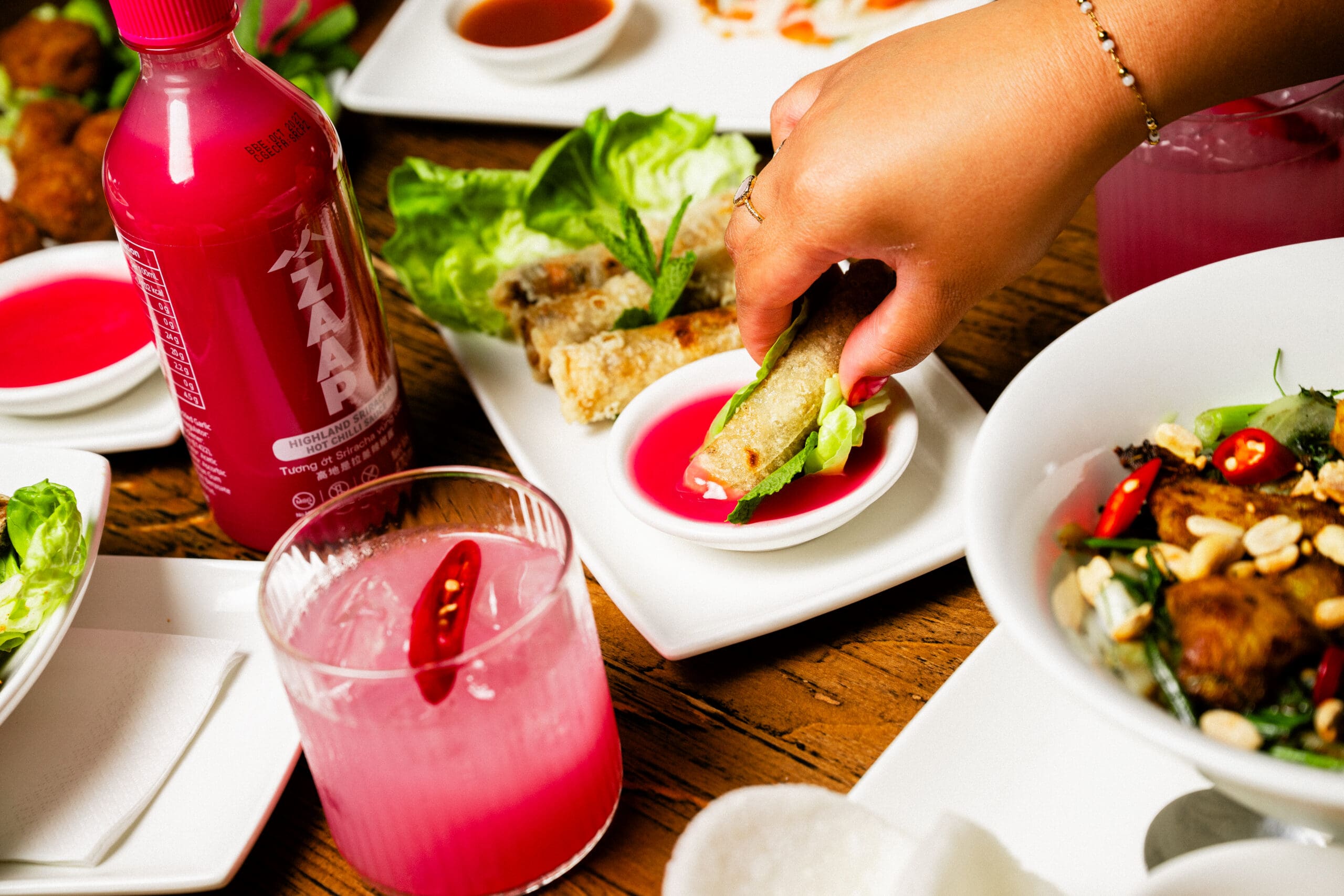 A hand dips a fresh spring roll into pink sauce, surrounded by a pink drink, bottle, salad, and assorted Asian dishes on a wooden table.