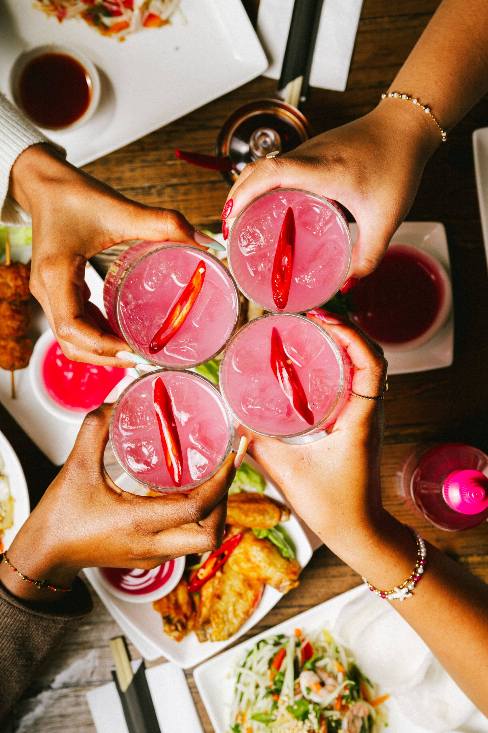 Four people clink pink cocktails garnished with red chili peppers over a table set with assorted Asian dishes.