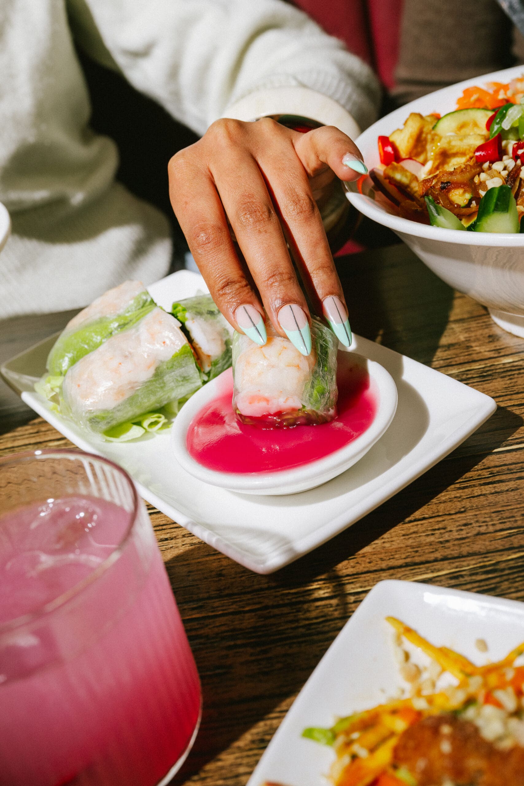 A hand with painted nails dips a shrimp spring roll into pink sauce. Other plates of food and a pink drink sit on the wooden table.