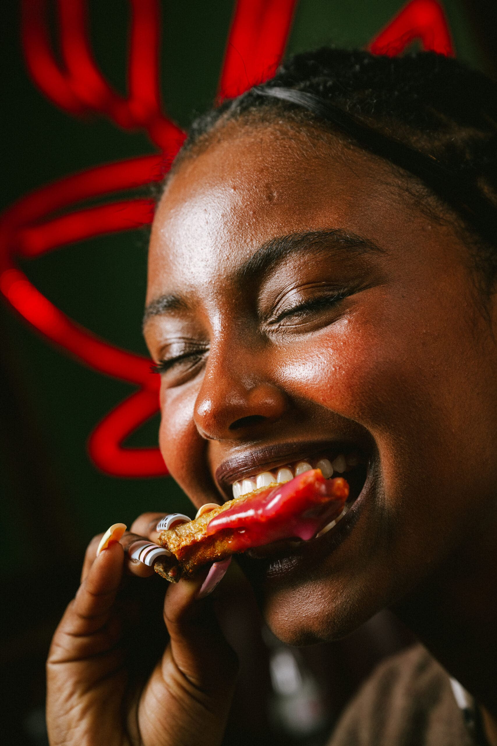 A person smiling and taking a bite of food with pink frosting or sauce, holding it close to their mouth. The background is blurred with red neon light shapes.