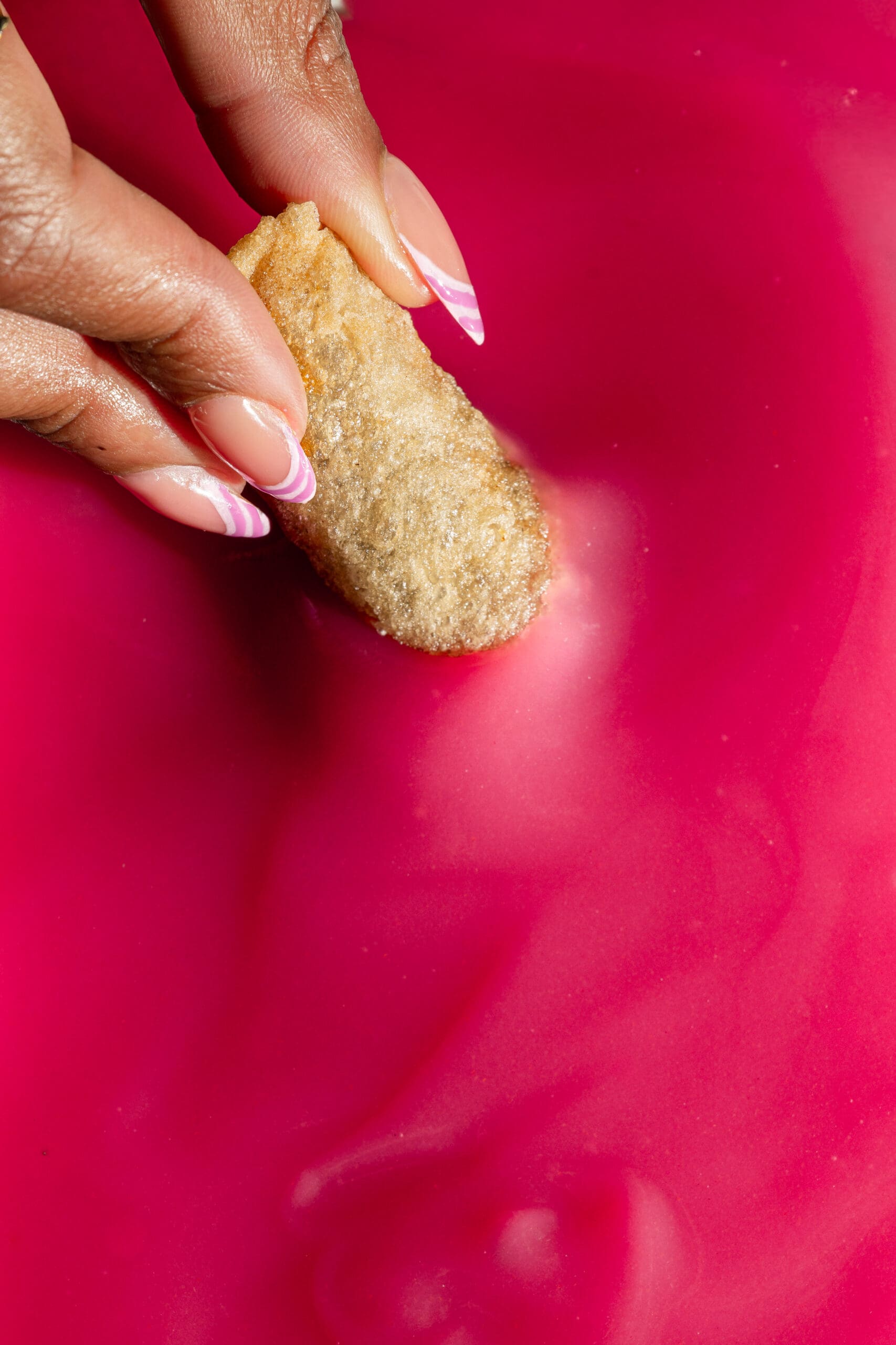 A hand with manicured nails dips a breaded food item into a thick, bright pink sauce or liquid.