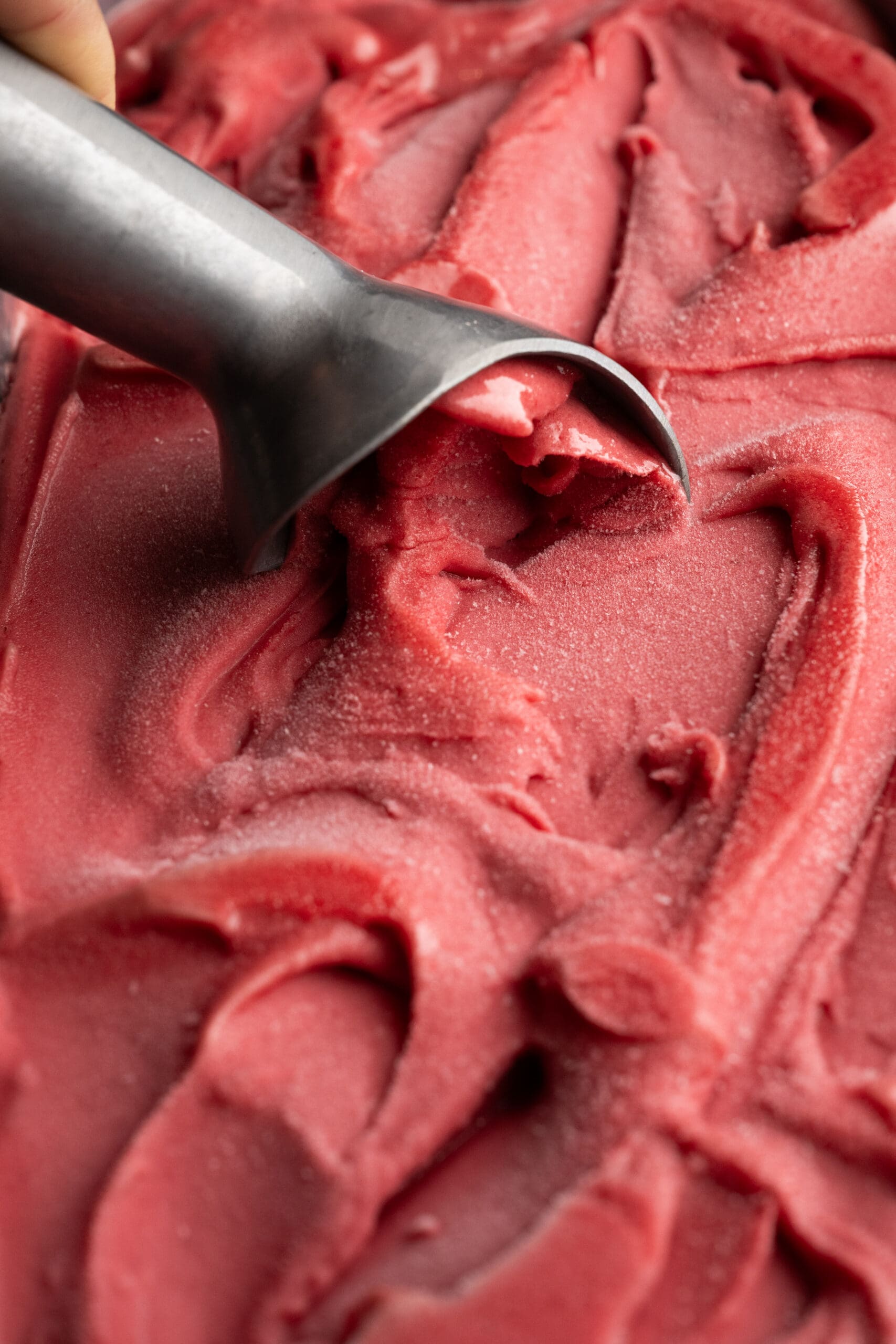 A close-up of a metal ice cream scoop gliding through smooth, red sorbet or ice cream, creating textured swirls on the surface.