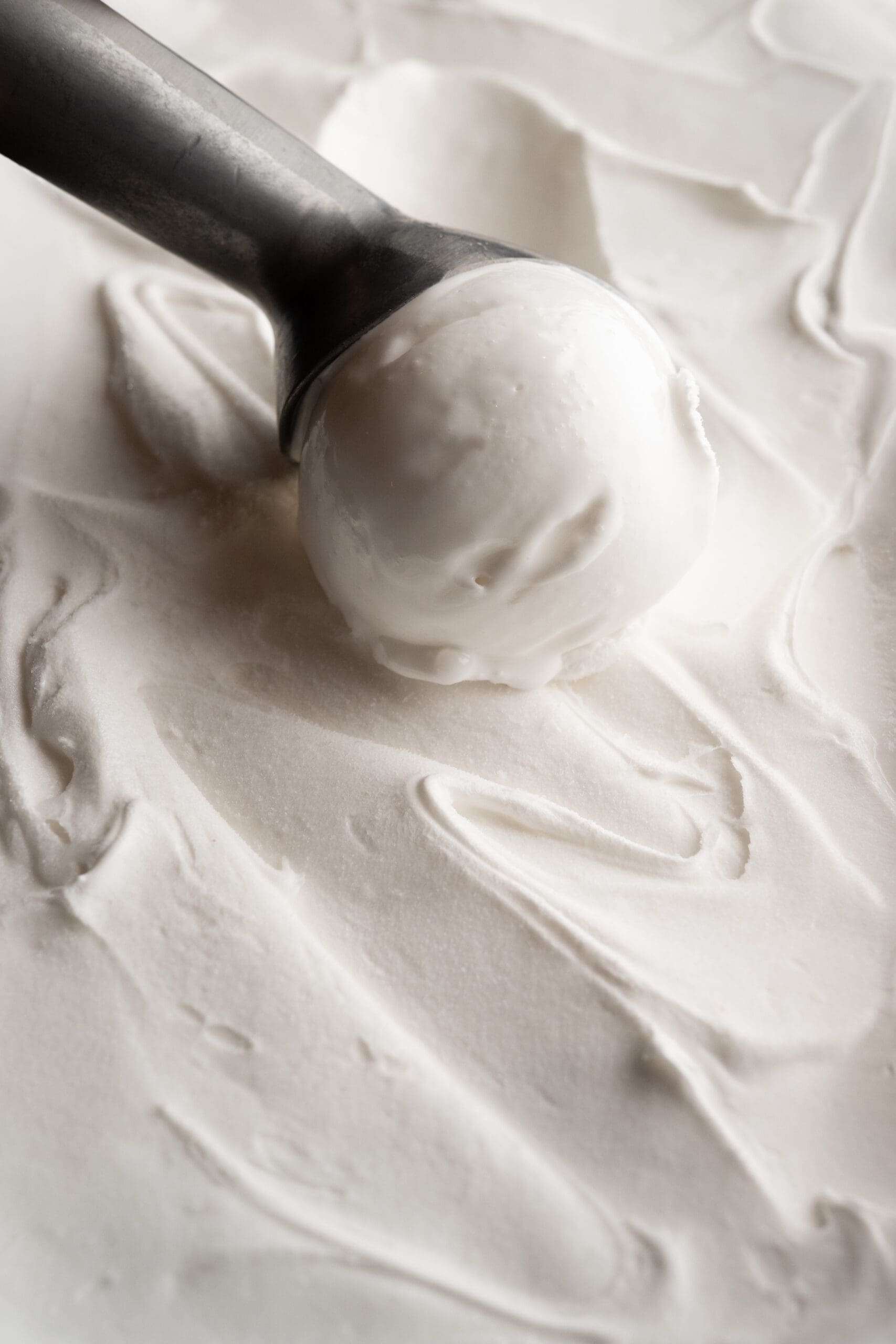 A metal ice cream scoop with a round scoop of vanilla ice cream rests on a smooth, creamy surface of more ice cream.